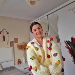 Puede incluir: Un c&aacute;rdigan de punto blanco con flores de ganchillo de colores. El c&aacute;rdigan tiene cierre de botones y mangas largas.