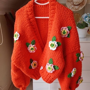 Può includere: Un cardigan di maglia arancione brillante con fiori all'uncinetto in tonalità di rosa, bianco, giallo e verde.