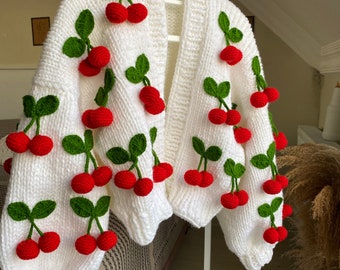Op de afbeelding: Witte gebreide cardigan met rode en groene kersen. De cardigan heeft een V-hals en lange mouwen.