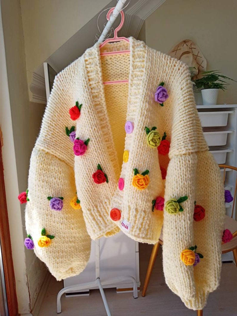Puede incluir: Un c&aacute;rdigan de punto amarillo p&aacute;lido con cierre de bot&oacute;n y adornos florales coloridos. El c&aacute;rdigan tiene un corte holgado y mangas largas.