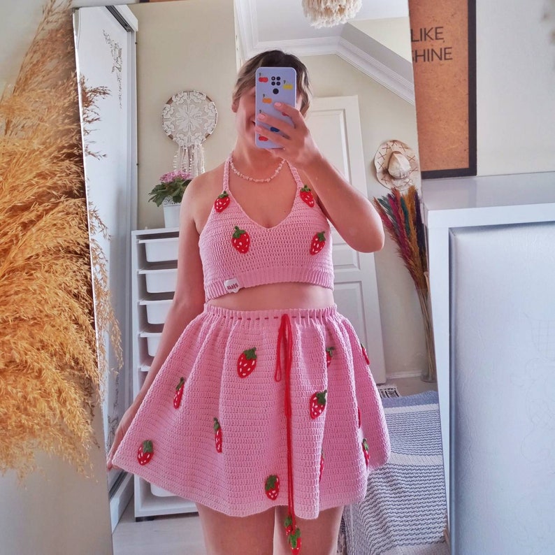 Pode incluir: Um conjunto de top halter e saia em croch&ecirc; rosa claro com um padr&atilde;o repetitivo de pequenas aplica&ccedil;&otilde;es de morangos vermelhos e verdes. A saia inclui um cord&atilde;o vermelho na cintura com detalhes de morango nas pontas e uma etiqueta branca.