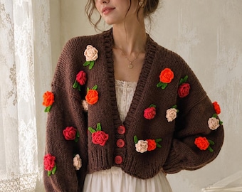May include: A dark brown knit cardigan featuring three red buttons and scattered three-dimensional crochet floral appliqués in shades of red, orange, and cream, each with small green leaves, worn over a white dress with lace detailing.