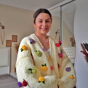 Puede incluir: Un c&aacute;rdigan de punto blanco con flores de ganchillo de colores. El c&aacute;rdigan tiene cierre de botones y mangas largas.