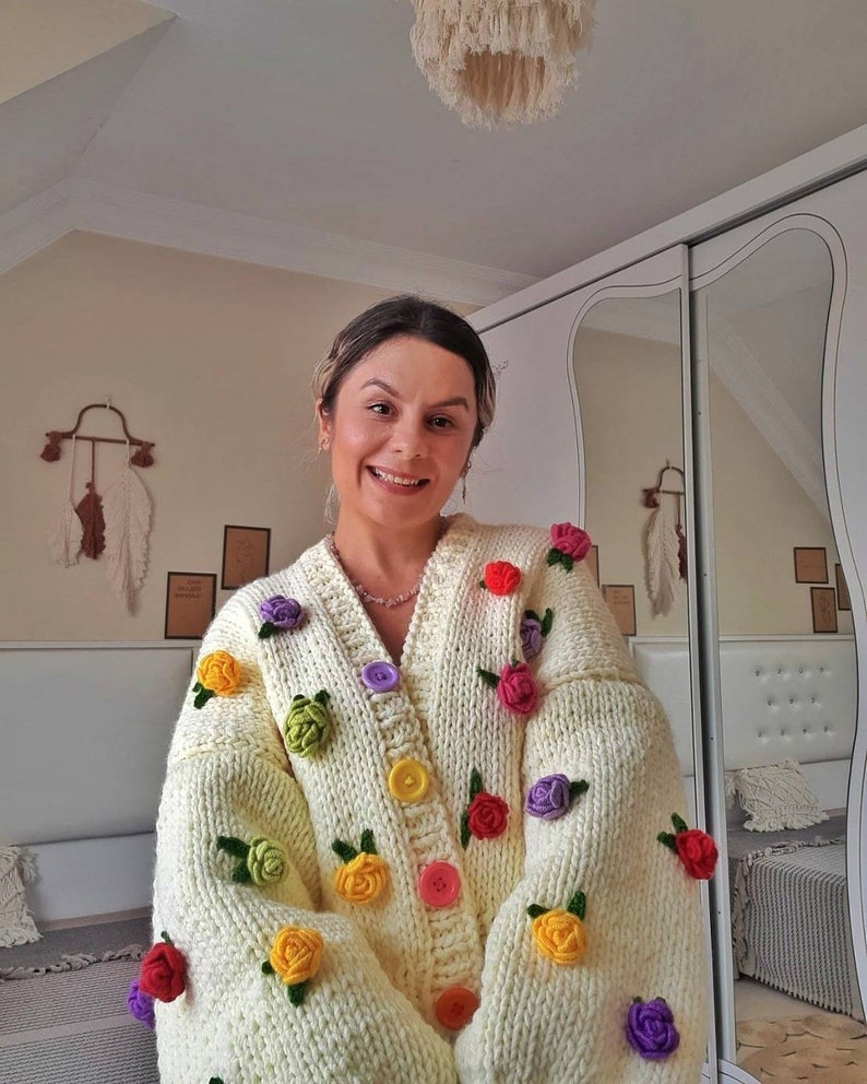 Puede incluir: Un c&aacute;rdigan blanco de punto con adornos de flores de ganchillo de colores. El c&aacute;rdigan tiene cierre de botones y mangas largas.