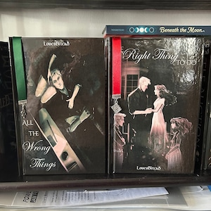 Puede incluir: Una estantería con cuatro libros, cada uno con una portada diferente. Los libros son todos negros con letras plateadas. El libro de la izquierda tiene el título "Manacled" y el libro de la derecha tiene el título "Beneath the Moon". Los dos libros del medio tienen el título "All the Wrong Things" y "Right Thing to Do". Cada libro tiene un marcapáginas con un diseño diferente.