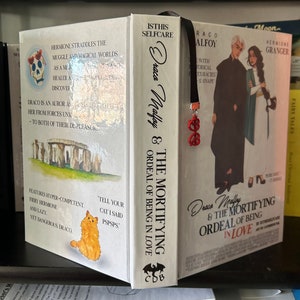 Könnte beinhalten: Ein weißes Buch mit dem Titel "Draco Malfoy & The Mortifying Ordeal of Being in Love" und einem schwarzen Lesezeichen mit einem roten Brillen-Anhänger. Der Buchumschlag zeigt eine schwarze Fledermaus mit den Buchstaben "CDB" auf ihren Flügeln. Das Buch befindet sich in einem Regal mit anderen Büchern.