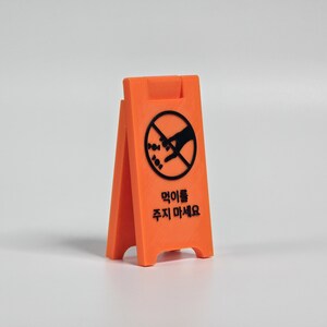 Könnte beinhalten: Ein orangefarbenes A-Rahmen-Schild mit schwarzem Text und einer Grafik einer Hand, die nach Futter greift, in einem durchgestrichenen Kreis. Auf dem Schild steht "Nicht füttern" auf Koreanisch.