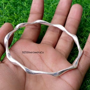 Puede incluir: Un brazalete de plata pulida con un diseño ondulado y continuo. La superficie metálica es lisa y reflectante, presentando un patrón curvo repetitivo en toda la circunferencia. El texto 925SilverJewelryCo es visible.