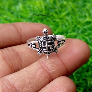 Puede incluir: Un anillo de tono plateado que presenta una pieza central con forma de tortuga y un diseño de caparazón detallado. El caparazón muestra un símbolo geométrico central de cuatro brazos. El aro tiene recortes decorativos y acentos oxidados.