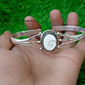 Peut inclure: Un bracelet manchette en argent avec une coquille d'œil de Shiva blanche ovale au centre. La coquille est encadrée par un bord en argent torsadé et montée sur une manchette à trois bandes. Le texte 925SilverJewelryCo est visible en haut à gauche.