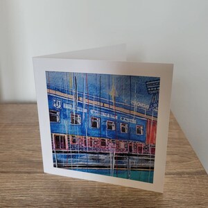 May include: A blue and red illustrated greeting card featuring a building with the words "The Stadium" written on the side. The card is on a wooden surface.