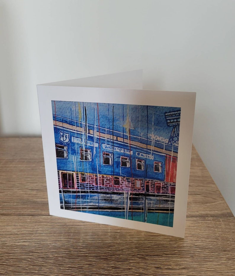 May include: A blue and red illustrated greeting card featuring a building with the words "The Stadium" written on the side. The card is on a wooden surface.