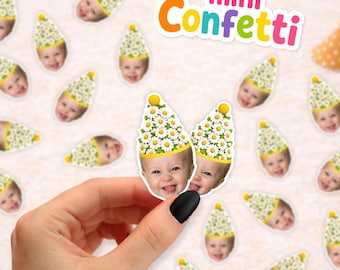 May include: Small paper confetti pieces featuring a smiling baby wearing a party hat decorated with white daisy flowers. The confetti is scattered across the surface, and the text mini Confetti is printed in colorful letters at the top.