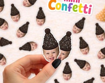 May include: Mini Confetti paper cutouts featuring a baby's face wearing a party hat made of chocolate sandwich cookies. The confetti pieces are scattered, and one piece is held up. There are also small yellow and blue polka-dot party hats.