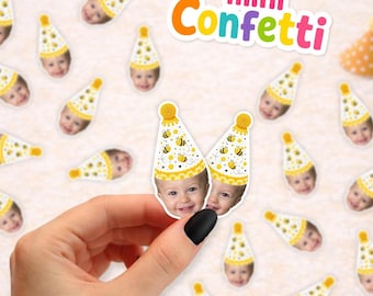 May include: Small paper confetti pieces featuring a photograph of a child wearing a yellow and white bee-patterned party hat. The text mini Confetti is displayed in colorful letters at the top. A hand holds two of the confetti pieces.