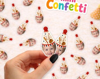 May include: Small paper confetti pieces featuring a child's face wearing a party hat decorated with baseballs, bats, and gloves. The confetti is scattered across a surface, with one piece held up to show the detailed print and red pom-pom topper.