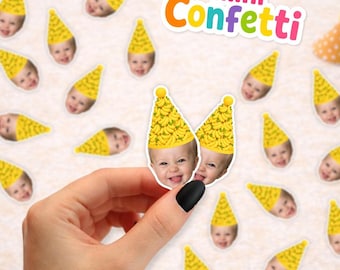 May include: Customized paper confetti featuring a repeating photo of a baby wearing a yellow party hat with a banana pattern. The confetti pieces are scattered across a surface, with one piece held up to show the detail of the baby's face.