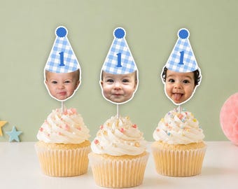May include: Three cupcakes topped with white frosting and sprinkles, each featuring a personalized cake topper. The toppers consist of a blue and white gingham party hat with the number 1, attached to a photo of a baby's face on a stick.