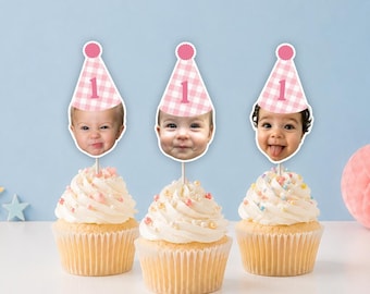 May include: Three cupcakes topped with white frosting and sprinkles, each featuring a personalized paper topper. The toppers display a photograph of a child's face wearing a pink and white gingham party hat with the number one printed on it.