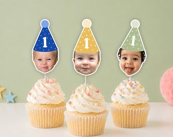 May include: Three cupcakes topped with personalized cake toppers featuring a child's face and a party hat with the number 1. The hats are blue, yellow, and light green with white polka dots. Each topper is attached to a thin white stick.