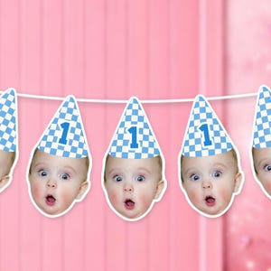 Puede incluir: Una guirnalda de fiesta con recortes repetidos de la cara de un bebé que lleva un gorro de fiesta a cuadros azules y blancos con el número uno impreso en el frente. Los recortes están unidos por un cordel blanco sobre fondo rosa.