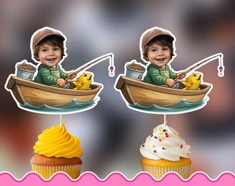 May include: Two printable cupcake toppers featuring a cartoon child in a brown cap and green shirt sitting in a wooden boat with a fishing rod, a bucket, and a yellow fish. The toppers are displayed on yellow and white frosted cupcakes.