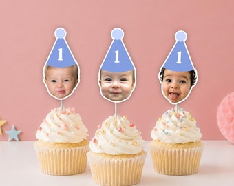 May include: Three cupcakes with white frosting and colorful sprinkles, each topped with a custom paper cutout of a baby's face wearing a light blue party hat with a white pom-pom and the number one printed in white on the front of the hat.