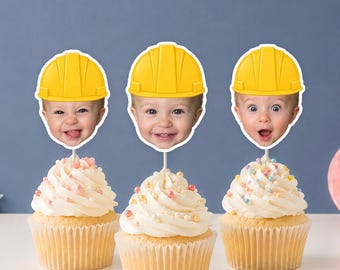 May include: Three cupcakes topped with white frosting and colorful sprinkles, each featuring a cutout of a baby's face wearing a bright yellow construction hard hat. The cutouts are attached to thin white sticks inserted into the center of each cupcake.