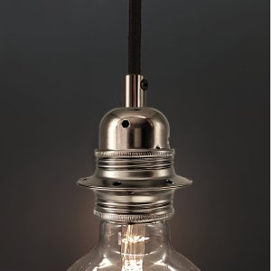 Peut inclure: Un luminaire en métal argenté avec un cordon noir. Le luminaire a un sommet rond et en forme de dôme avec un petit trou au centre. Le bas du luminaire a une base large et plate avec une série de crêtes autour du bord.