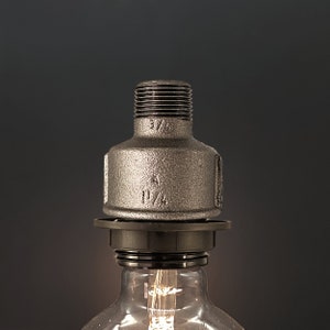 Op de afbeelding: Een zilveren metalen pijpfitting met de markeringen "3/4" en "1 1/4" bevestigd aan een zwarte metalen lampfitting.