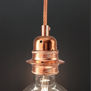 Op de afbeelding: Een koperkleurige lamp met een gevlochten snoer en een ronde, decoratieve voet. De lamp is ontworpen om een ​​lichtbol te houden.