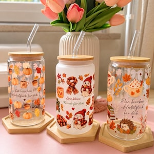 Puede incluir: Tres vasos de cristal con tapas de bambú y pajitas de vidrio, cada uno con ilustraciones de temática otoñal de calabazas, hojas y motivos acogedores, con texto en alemán que incluye 'Ein bisschen Herbstglück für dich' y 'Eine kleine Freude für dich'.