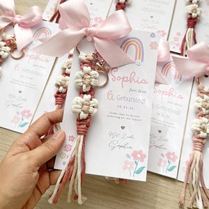 May include: A macrame keyring featuring two cream-coloured flower knots and a dusty rose cord with a tassel, attached to a card printed with a rainbow, the name Sophia, and the text 6. Geburtstag. A light pink satin ribbon bow is tied at the top.