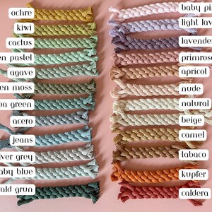 May include: A collection of twenty-two braided cotton cords arranged in two vertical columns. Each cord is labelled with colour names including ochre, kiwi, cactus, green pastel, agave, green moss, spa green, acero, jeans, silver grey, baby blue, wald grun, baby pink, light lavender, lavender, primrose, apricot, nude, natural, beige, camel, tabaco, kupfer, and caldera.