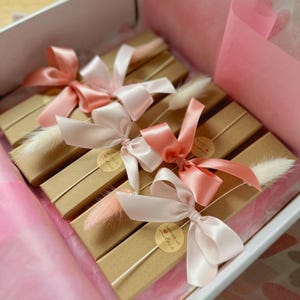 May include: Four rectangular brown cardboard boxes arranged in a white gift box. Each box is tied with a satin ribbon in shades of pink and peach, accented with a small dried white or pink grass plume and a round sticker reading handmade with love.