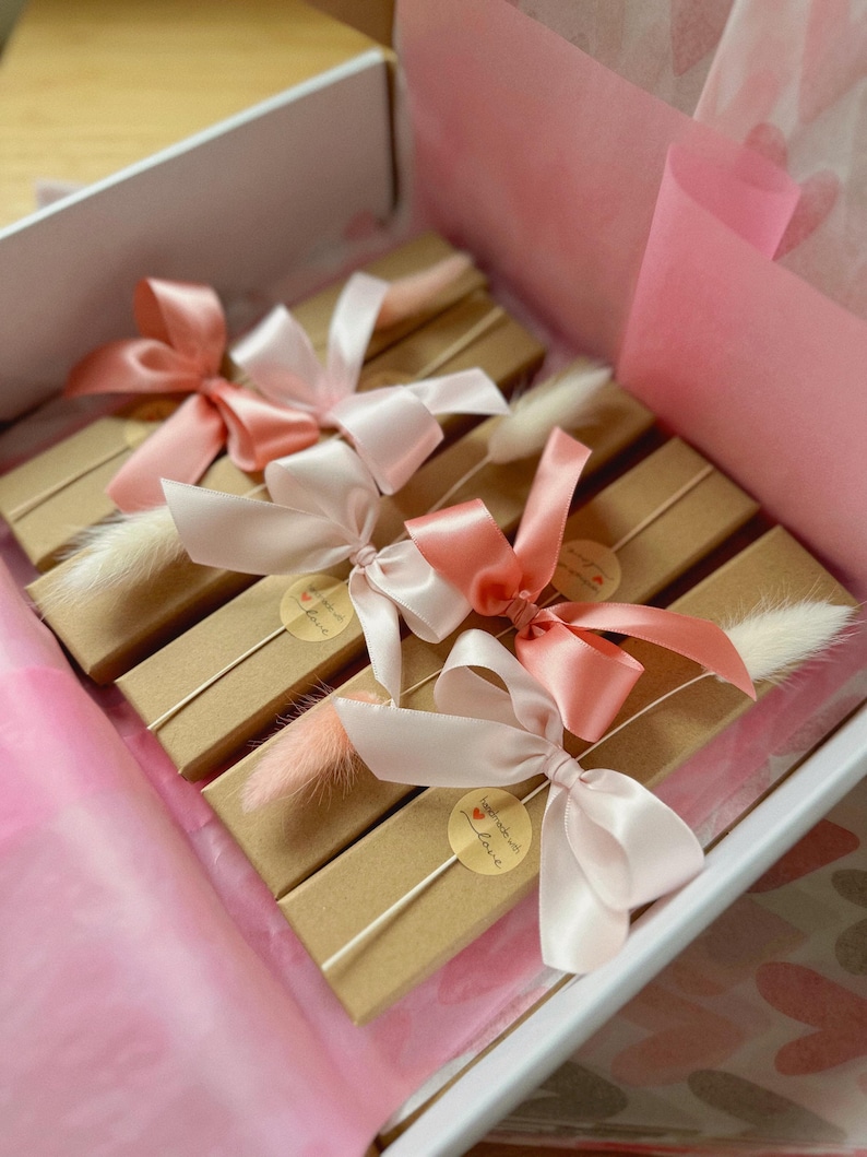 May include: Four rectangular brown cardboard boxes arranged in a white gift box. Each box is tied with a satin ribbon in shades of pink and peach, accented with a small dried white or pink grass plume and a round sticker reading handmade with love.
