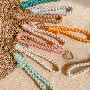 May include: A collection of seven macrame wristlet keychains in various colours including teal, light grey, tan, mustard yellow, dusty pink, cream, and mint green. Each keychain features a braided cord loop and a gold-toned heart-shaped metal clasp.