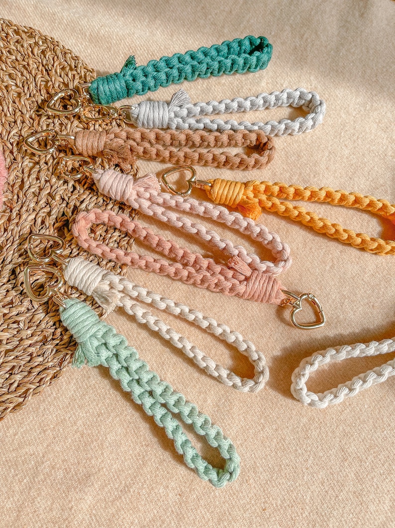 May include: A collection of seven macrame wristlet keychains in various colours including teal, light grey, tan, mustard yellow, dusty pink, cream, and mint green. Each keychain features a braided cord loop and a gold-toned heart-shaped metal clasp.