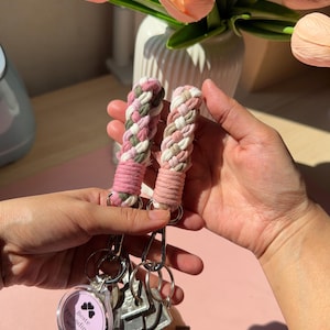 Peut inclure: Deux porte-clés en macramé tressé composés de cordons en coton rose, blanc et vert. L'un des porte-clés comporte une breloque ronde avec le texte "Beste Freundinnen seit 2019" et une icône de cœur. Ils sont fixés à des mousquetons et anneaux en métal argenté.