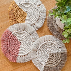 Könnte beinhalten: Vier handgefertigte Makramee-Untersetzer in Weiß, Beige, Rosa und Grau. Jeder Untersetzer hat ein Spiraldesign mit Fransen. Die Untersetzer sind auf einer Holzoberfläche angeordnet, mit einer kleinen Pflanze im Hintergrund.