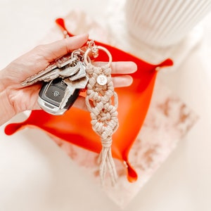 May include: A beige macrame keyring featuring a gold-toned metal ring and a small circular charm with the letter P engraved on it. The keyring is attached to a set of metal keys and a black car key fob, showcasing intricate knotted cord patterns.