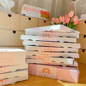 May include: A stack of white cardboard shipping boxes with light pink belly bands featuring the text "MODELA" and a rainbow graphic. The boxes are arranged in a vertical pile, with additional boxes placed to the left on a wooden surface.