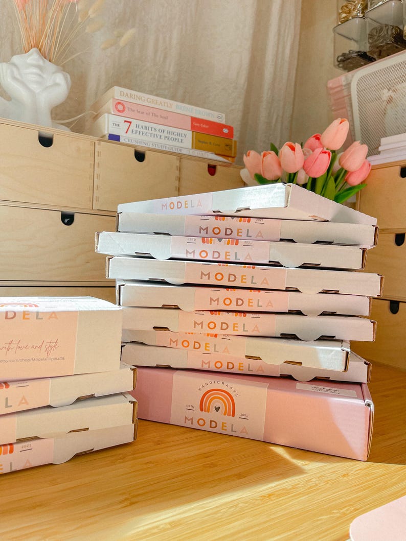 May include: A stack of white cardboard shipping boxes with light pink belly bands featuring the text "MODELA" and a rainbow graphic. The boxes are arranged in a vertical pile, with additional boxes placed to the left on a wooden surface.