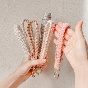 Könnte beinhalten: Ein Set aus fünf Makramee-Schlüsselbändern in den Farben Creme, Beige, Grau und Rosa. Jedes Schlüsselband besteht aus einer geflochtenen Kordel und verfügt über einen silberfarbenen Metallverschluss in Herzform zur Befestigung von Schlüsseln.