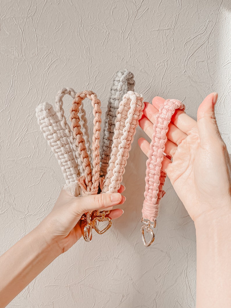May include: A collection of five macrame wristlet keychains in shades of cream, tan, grey, and pink. Each keychain features a braided cord design and a silver-toned metal clasp shaped like a heart at the base for attaching keys or accessories.
