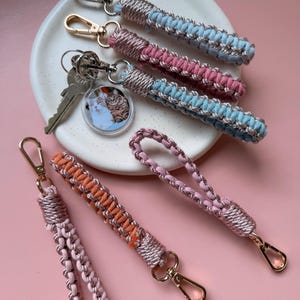 Pode incluir: Cinco porta-chaves de pulso em macramé com cordão metálico em ouro rosa entrelaçado com cordão de algodão azul pastel, rosa, laranja e rosa claro. Cada porta-chaves inclui um mosquetão dourado e um tem um pendente circular com foto de gatos.
