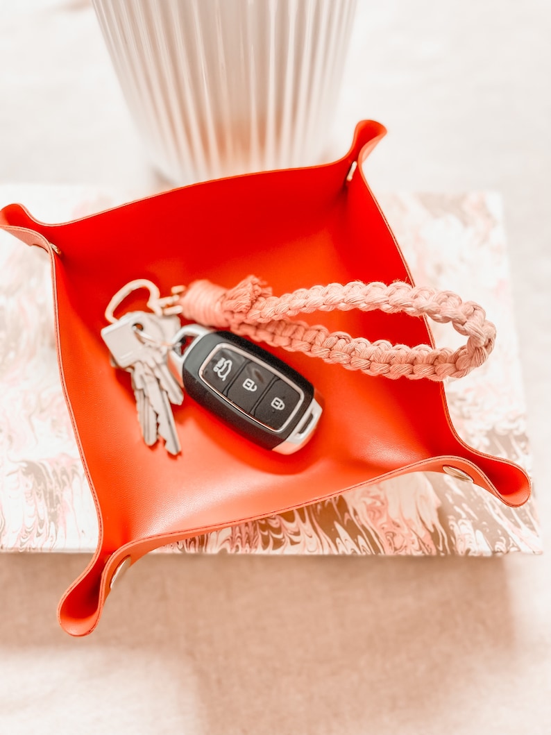 May include: A bright orange square valet tray with snap-button corners containing a set of metal keys with a black plastic remote fob and a light pink braided macrame wristlet strap attached to a gold-toned heart-shaped metal keyring.