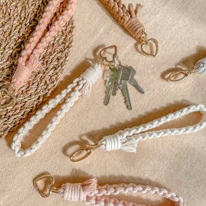 May include: A collection of macrame wristlet keychains in soft pink, beige, and cream colours. Each keychain features a braided cord, a gold-toned metal heart-shaped clasp, and a knotted tassel detail. One cream keychain holds a set of silver metal keys.