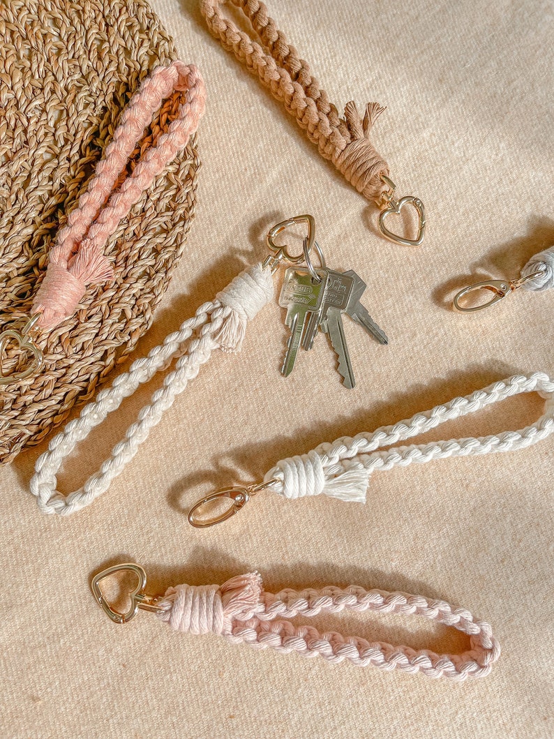 May include: A collection of macrame wristlet keychains in soft pink, beige, and cream colours. Each keychain features a braided cord, a gold-toned metal heart-shaped clasp, and a knotted tassel detail. One cream keychain holds a set of silver metal keys.