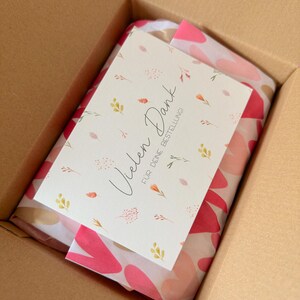 May include: A brown cardboard shipping box containing a package wrapped in white tissue paper with a pink and red heart pattern. A white rectangular card rests on top, featuring small floral illustrations and the text Vielen Dank F&Uuml;R DEINE BESTELLUNG.